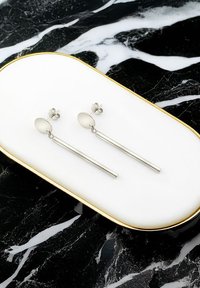 Silverfärgade hängörhängen med oval stift och avlånga raka stänger, visade på en vit bricka med guldkant. Texturerad yta synlig.