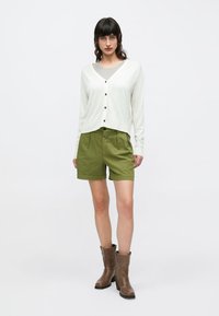 Ελαφρύ μπεζ ζακέτα με μακριά μανίκια και κουμπιά, συνδυασμένη με χακί σορτς. Το σύνολο ολοκληρώνεται με καφέ μποτάκια μέχρι τον αστράγαλο. Απλός σχεδιασμός συνολικά.