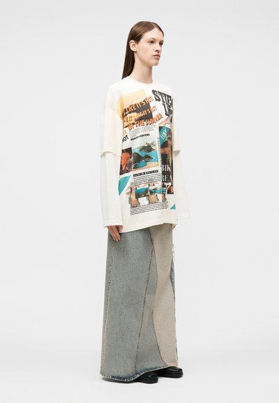 Νεαρή γυναίκα που φοράει λευκή μπλούζα με γραφικό σχέδιο και μακριά φούστα από τζιν με πατς, στέκεται σε απλό λευκό φόντο.