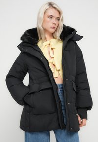 Svart polstret jakke med hette i imitert pels, glidelås foran, trykknapper og midjebelte. Teksturert stoff med tydelig quilting.