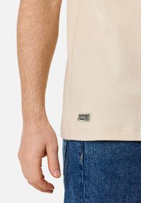 Beige cotton t-shirt with a straight cut, featuring a woven label on the bottom. Blue denim jeans with a slightly distressed texture.