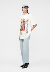 Frau trägt ein übergroßes weißes T-Shirt mit Grafik, hellblaue Jeans, schwarze spitze Stiefeletten und eine hellblaue Jeansmütze, steht mit einer Hand in der Tasche.