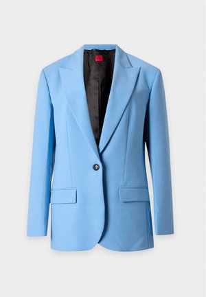 Femme aux longs cheveux noirs portant un costume bleu clair avec un pantalon large et une chemise blanche, debout sur un fond clair uni.