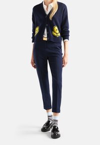 Mörkblå cardigan med gula tecknade applikationer, vit skjorta, mörkblå byxor, randiga strumpor och svarta loafers med kraftiga sulor.