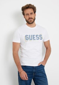 Balta medvilninė marškinėlių su trumpomis rankovėmis, kuriuose užrašyta žodis "GUESS" dideliais, išblukusiais mėlynais ir smėlio spalvos rašmenimis. Golfo formos apykaklė ir laisvas siluetas.