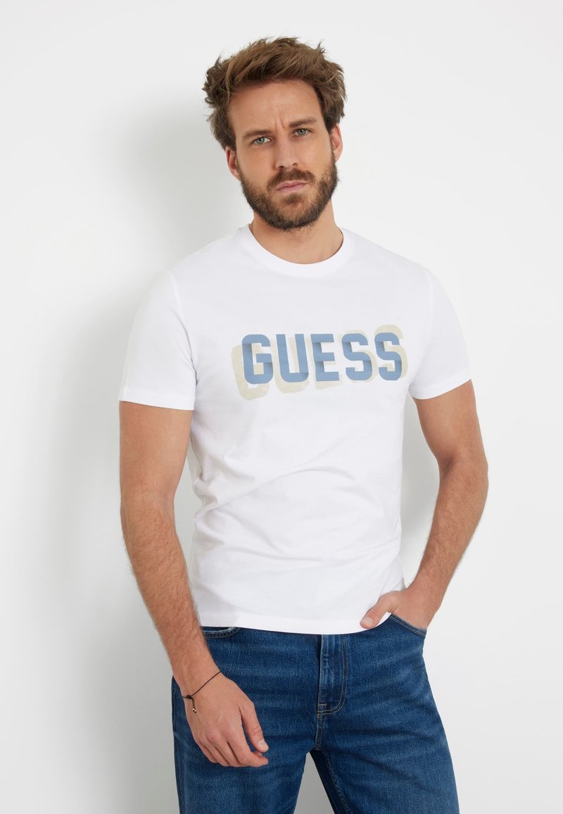 Balta medvilninė marškinėlių su trumpomis rankovėmis, kuriuose užrašyta žodis "GUESS" dideliais, išblukusiais mėlynais ir smėlio spalvos rašmenimis. Golfo formos apykaklė ir laisvas siluetas.