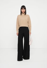 Pull cropped beige avec ourlet côtelé associé à un pantalon noir large. Le modèle tient un petit sac à main rose clair. Fond blanc uni.