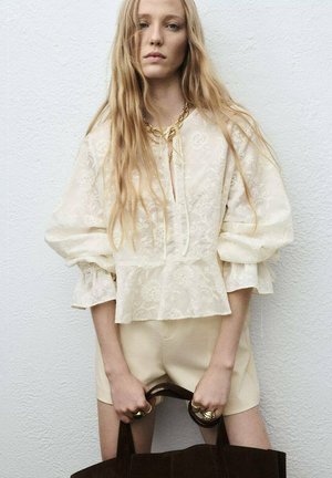Blouse crème brodée de fleurs avec des manches bouffantes, un ourlet peplum et un col à nouer. Associée à un short clair et un sac sombre. Accents de bijoux en or.