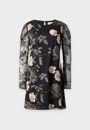 Femme aux cheveux bouclés portant une robe noire à fleurs avec des manches bouffantes transparentes et des sandales dorées à talons hauts, debout devant un fond uni.