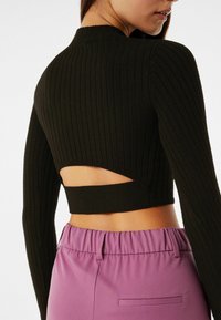 Kvinna i en ribbad svart långärmad crop top med ett snitt på ryggen, som är kombinerad med hög midja lila byxor med resårmidja.