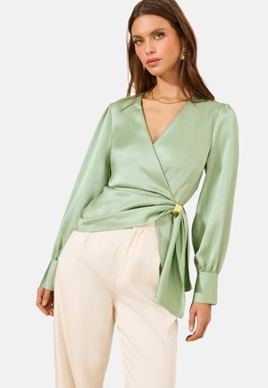 Frau trägt eine hellgrüne Satin-Wickelbluse mit langen Puffärmeln und cremefarbenen hochtaillierten Hosen vor einfachem Hintergrund.