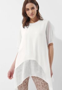 Witte tuniek top met korte, semi-doorzichtige mouwen, een ronde halslijn en een langere, gebogen zoom, gelaagd over patroonrijke, lichtbruine broek.