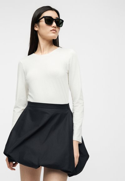 Femme portant des lunettes de soleil noires, un haut blanc à manches longues et une jupe noire volumineuse, debout devant un fond uni.