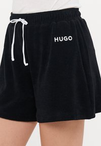 Svarta velourshorts med elastisk midja, vit dragsko och broderad "HUGO"-logotyp på vänstra låret. Mjuk textur.