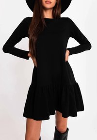 Czarna sukienka z długim rękawem i falbaną na dole, wykonana z miękkiego materiału o gładkiej fakturze. Posiada luźny krój i okrągły dekolt.