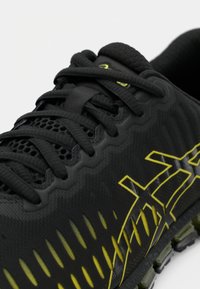 Gros plan d'une chaussure de sport noire avec tige en mesh, lacets noirs et logo et design de marque jaunes sur le côté.