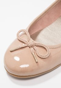 Ballerine beige brillante avec nœud tressé et extrémités dorées sur une semelle intérieure texturée claire et une semelle souple.