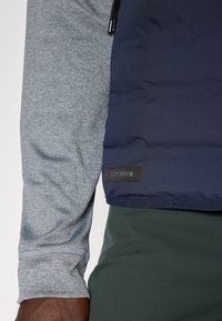 Gilet imbottito blu navy con finitura opaca e maniche lunghe grigie sotto. Presenta una toppa di branding nera e un cordoncino sul colletto.