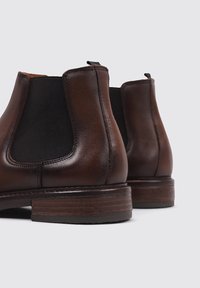 Bottes Chelsea en cuir marron avec des côtés élastiques noirs, design courbé et talon en bois empilé. Texture lisse avec une finition brillante.