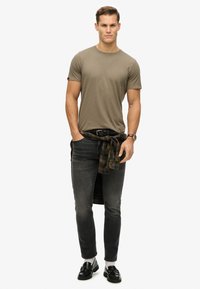 Olijfgroene t-shirt, slim fit jeans, geruit shirt om de taille geknoopt, zwarte instappers en grijze sokken. Eenvoudig ontwerp met minimale accenten.
