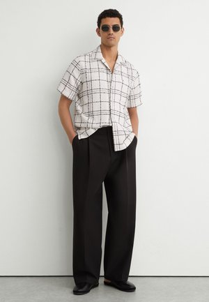 Homme portant des lunettes de soleil, chemise blanche à manches courtes avec motif quadrillé noir, pantalon large noir et chaussures noires, debout les mains dans les poches.