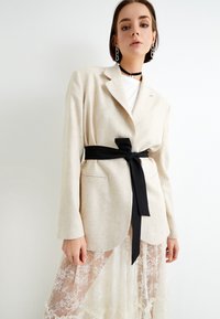 Beige linneblazer med ett svart tygbälte, kombinerad med en vit topp och en genomskinlig spetskjol med intrikata blommönster.