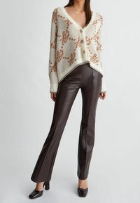 Cardigan de punto crema con patrón floral naranja, escote en V y tres botones. Pantalones de imitación de cuero marrón con corte recto y acabado brillante.