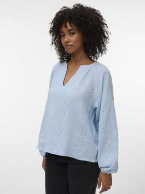 Göndör hajú nő, aki laza, világoskék, hímzett lyukacsos blúzt és fekete nadrágot visel, egyszínű fehér háttér előtt áll.