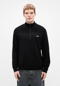 Μαύρο πουλόβερ με φερμουάρ στη ζώνη του λαιμού και υφή στο επάνω μέρος, μαλακό ύφασμα και μικρό λευκό λογότυπο στο στήθος. Μακριά μανίκια και ριγέ μανσέτες.