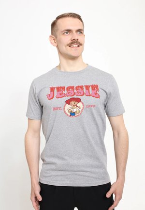 Mand med kort hår og overskæg står smilende, iført en grå T-shirt med Jessie-tegneserie, rød tekst og "EST 1999" trykt foran.