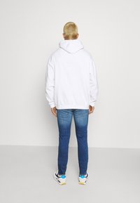 Weißer Kapuzenpullover, lockere Passform, mit gerippten Bündchen und Saum. Getragen mit blauen Skinny-Jeans und weißen Sneakern mit bunten Akzenten.