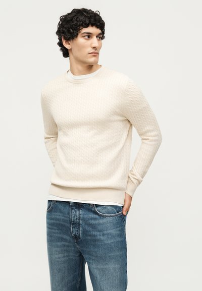 Άνδρας με σγουρά μαλλιά φορώντας ένα κρεμ πουλόβερ με πλεξούδα πάνω από ένα λευκό πουκάμισο και μπλε τζιν, κοιτάζοντας στο πλάι με τα χέρια πίσω από την πλάτη.