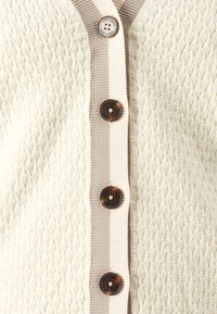 Cream textured cardigan with a ribbed beige placket and five brown buttons featuring a logo design. The texture is woven and subtle.