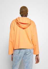 Huppari vaalean oranssista puuvillasta, vajaamittainen malli leveillä hihoilla, jossa on kengurupocket ja pehmeä, rakenteellinen pinta.