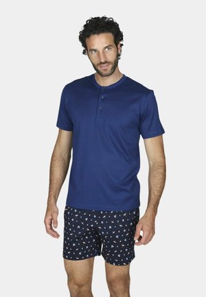 Uomo con capelli ricci che indossa una maglietta henley a maniche corte blu navy e pantaloncini blu navy con un piccolo motivo colorato.