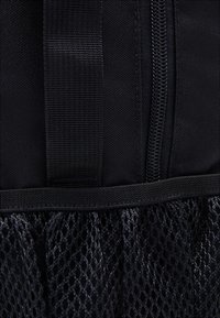 Gros plan d’un sac à dos en tissu noir montrant la fermeture éclair, la sangle solide et la poche en filet en bas.