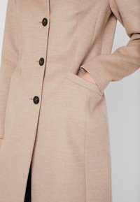 Lys beige frakke lavet af et blødt, struktureret stof, med sorte knapper, en enkelt brystlomme og sidelommer. Lange ærmer med manchetter.