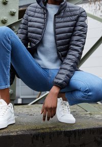 Sort dunjakke med quiltet design, lysegrå sweater, blå jeans og hvide sneakers med guld accenter, poseret mod industriel baggrund.