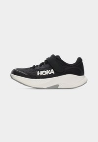 Αθλητικό παπούτσι HOKA ασπρόμαυρο με λουράκι Velcro και κορδόνι, ανώτερο μέρος από διαπνέον δίχτυ και επενδυμένη σόλα, πλάγια όψη σε λευκό φόντο.