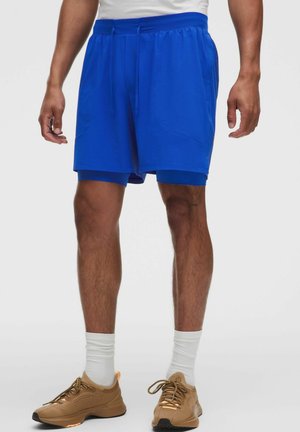 Mand iført lyseblå dobbel-lag atletikshorts, hvide sokker og brune sneakers, stående mod en ensfarvet baggrund.