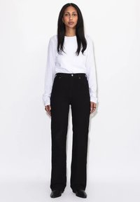 Frau mit langen dunklen Haaren, die vor einem weißen Hintergrund steht, trägt ein weißes Langarmshirt, schwarze hochtaillierte Hose und schwarze Schuhe.