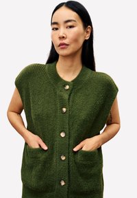 Vrouw met lang zwart haar, gekleed in een groen gebreid vest met knopen en zakken, met haar handen erin gestoken, staande tegen een witte achtergrond.
