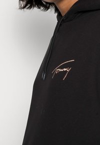 Sudadera negra con un tejido texturizado, que presenta un logo en cursiva color rosa dorado en el pecho y una capucha con cordón.