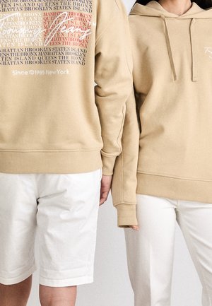 Zwei Personen tragen beige Kapuzenpullover und weiße Unterteile, eine in Shorts und die andere in Hosen, stehen nebeneinander vor einem schlichten Hintergrund.