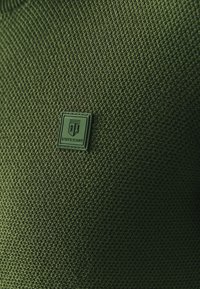 Donkergroen gewatteerde stof met een vierkante badge waarin "STATE of ART" is geëmbossed. Het materiaal lijkt geweven en duurzaam.