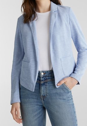 Kobieta w jasnoniebieskiej marynarce na białej koszuli i niebieskich dżinsach, z jedną ręką w kieszeni, a drugą swobodnie ułożoną wzdłuż ciała.