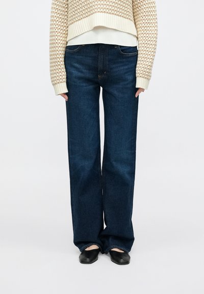 Tmavomodré džínsy s širokými nohavicami, ktoré majú hladkú denimovú textúru, vysoký pás a klasický päťvreckový dizajn. Nosené s béžovým pleteným svetrom.