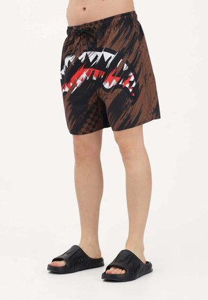 Parte inferior del cuerpo de una persona que lleva sandalias negras y pantalones cortos marrones con patrones abstractos en rojo, blanco y negro.