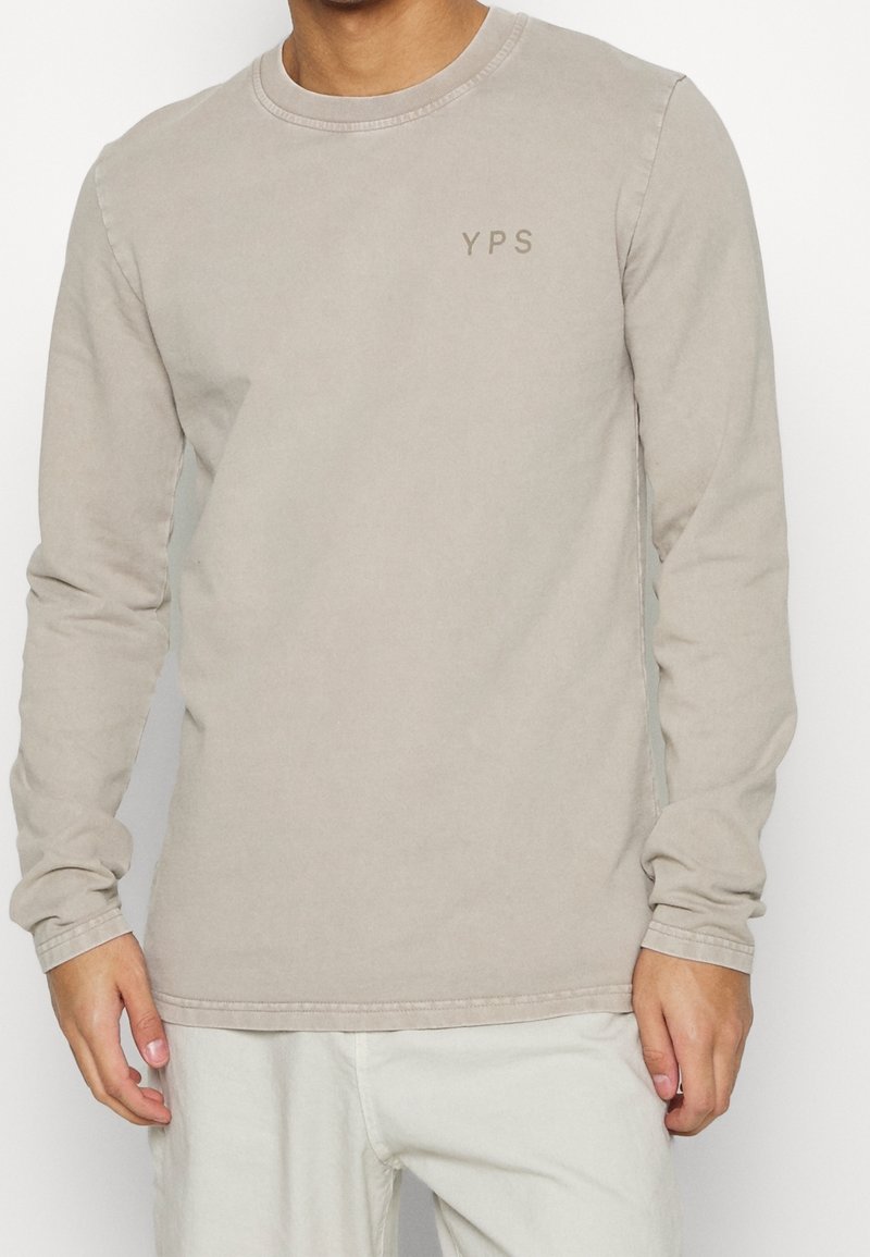 Košile s dlouhým rukávem z lehké béžové bavlny. Má kulatý výstřih a nenápadné logo "YPS" v mírně tmavším odstínu. Hladká textura, ležérní střih.