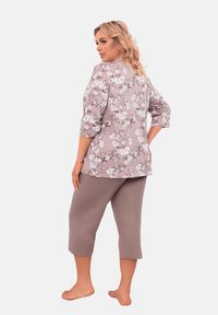 Kobieta stojąca boso, ubrana w fioletową, kwiatową bluzkę z trzy czwarte rękawem oraz dopasowane fioletowe spodnie typu cropped, lekko zwrócona bokiem z ręką na biodrze.